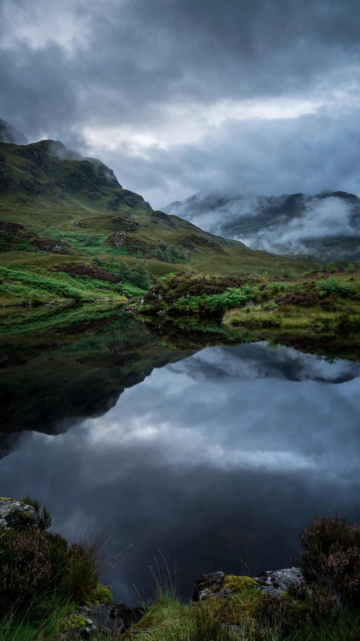 Scottish highlands with a perfectly still dark loch reflecting moody clouds and green hills