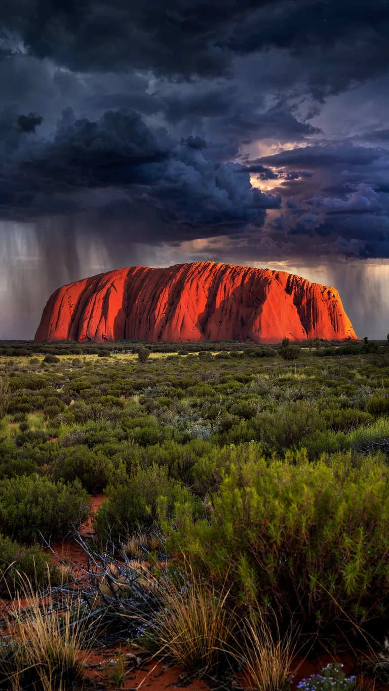 Uluru monolith glowing red at dusk under storm clouds, vertical landscape wallpaper