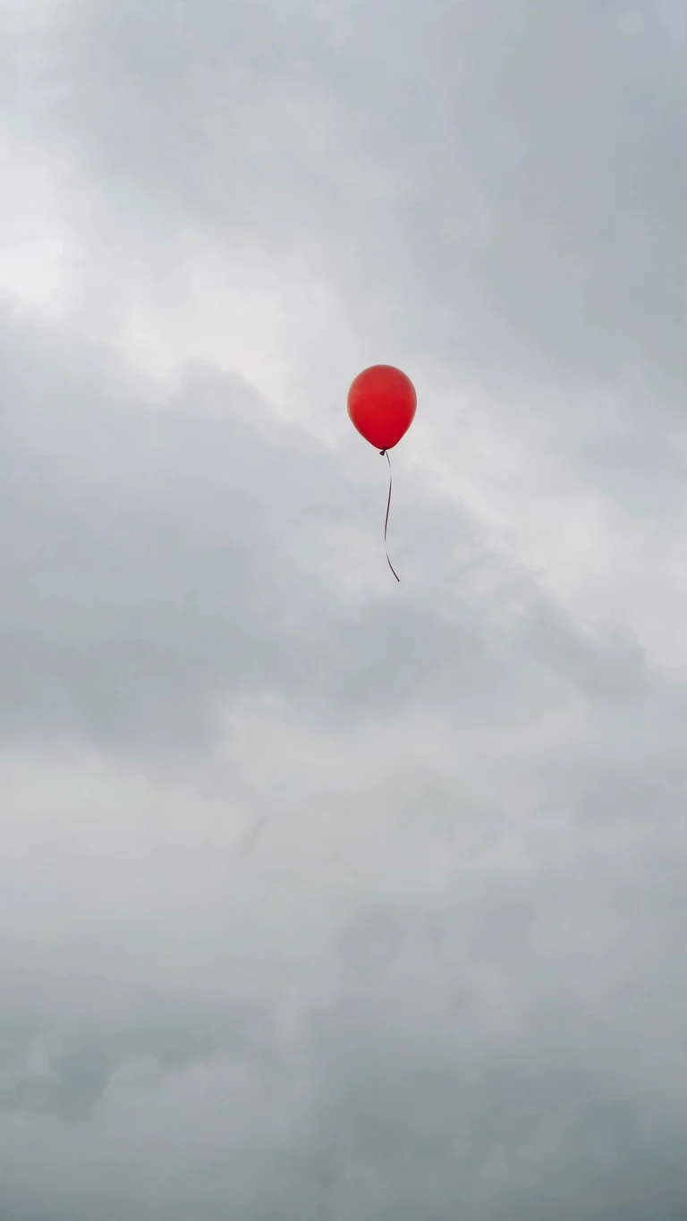 Single bright red balloon floating in a soft gray cloudy sky